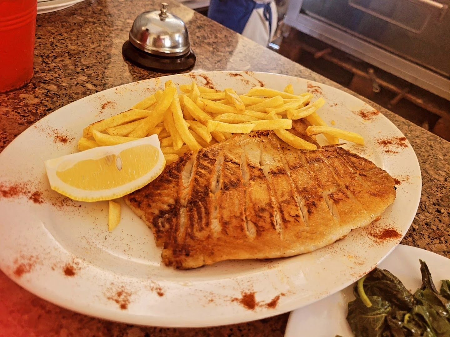 A whole grilled fish fillet with char marks, served with fries and a lemon wedge